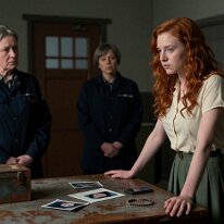 The Expropriation – Surrender of Identity A young woman at the counter of a decayed, dystopian neo-reformatory prison for women, placing her hands on a worn wooden desk with personal items, observed by...