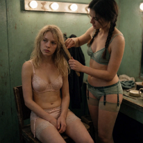 A Fleeting Moment of Tenderness Backstage In a cramped backstage room, a young brunette carefully brushes the tangled hair of a seated blonde. Both wear undergarments and garter belts. Her patient,...