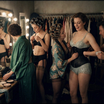 Grim Sisterhood in a Cramped Dressing Room Two young women share a tiny backstage dressing room. One in a greyish bra with lace trim struggles with loose seamed stockings. The other, in a black satin...